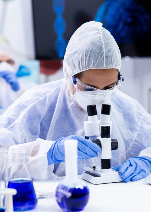 Female scientist looking through a microscope in research laboratory. Smoking blue liquid in test tubes.